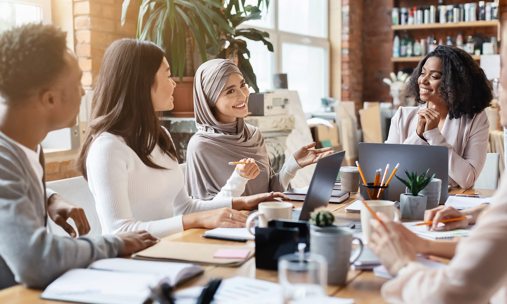 Ein multikulturelles Team diskutiert fröhlich in einem modernen Büro mit Laptops und Pflanzen auf dem Tisch.